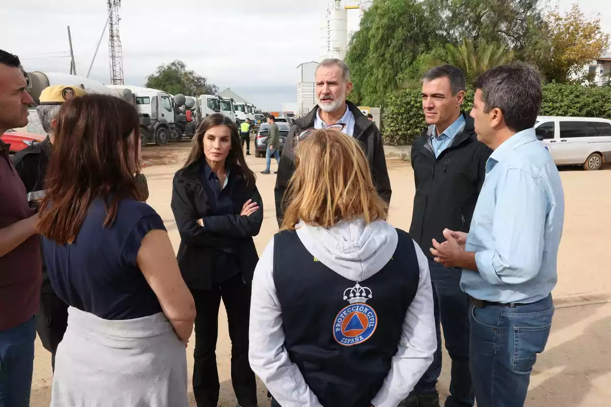 Un grupo de personas conversando al aire libre, algunas llevan chaquetas y una persona tiene un chaleco de Protección Civil de España, con camiones y vegetación al fondo.