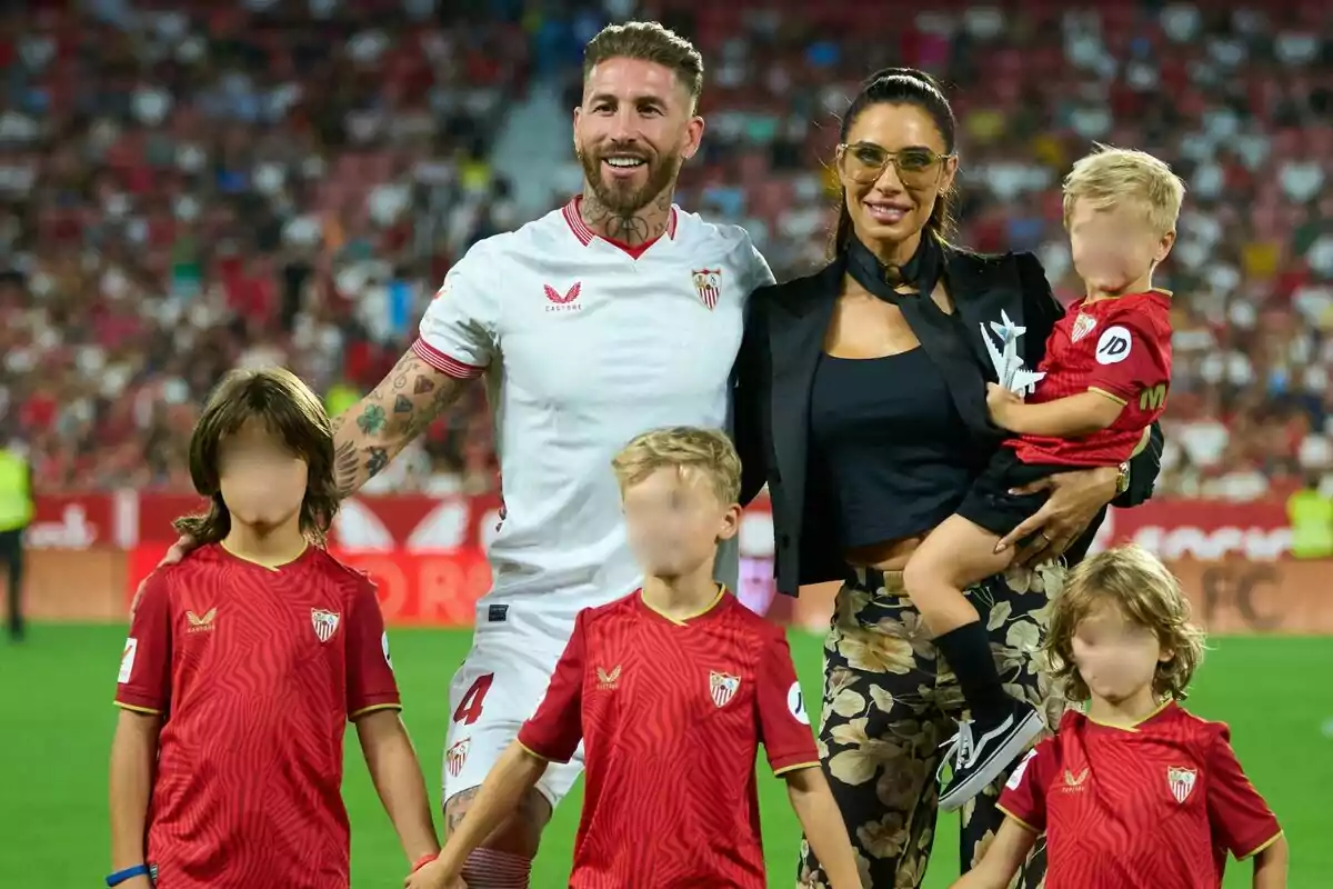 Un jugador de fútbol con uniforme blanco del Sevilla FC está en el campo de juego acompañado de una mujer y cuatro niños, todos con camisetas rojas del equipo, en un estadio lleno de espectadores.