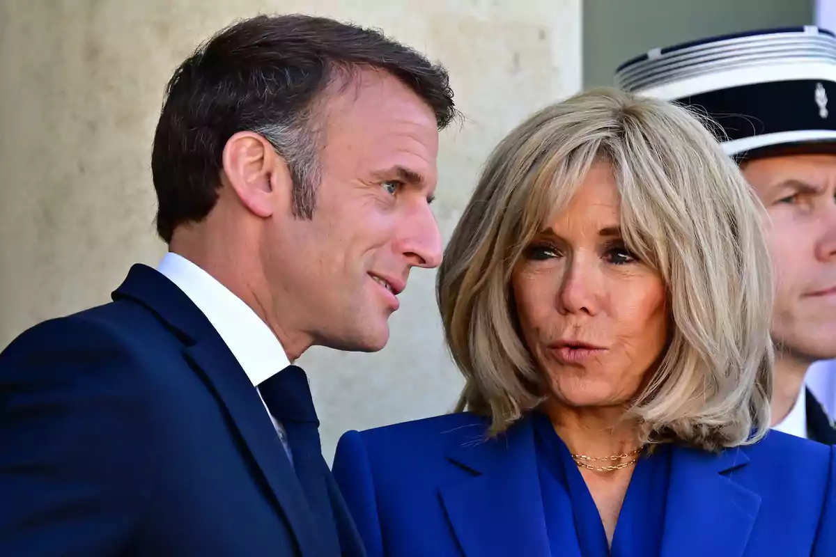 Emmanuel Macron i Brigitte Macron vestits de manera elegant conversen mentre un guàrdia amb uniforme apareix al fons. Emmanuel Macron i Brigitte Macron vestits de manera elegant conversen mentre un guàrdia amb uniforme apareix al fons.