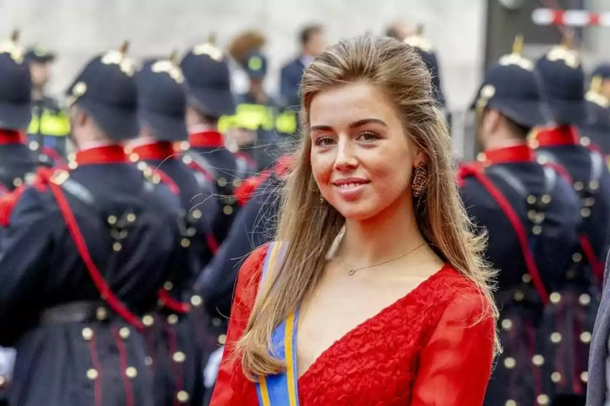 Alexia d'Holanda amb un vestit vermell i una banda blava i groga, somrient davant d'un grup de persones uniformades amb vestits foscos i barrets. Alexia d'Holanda amb un vestit vermell i una banda blava i groga, somrient davant d'un grup de persones uniformades amb vestits foscos i barrets.