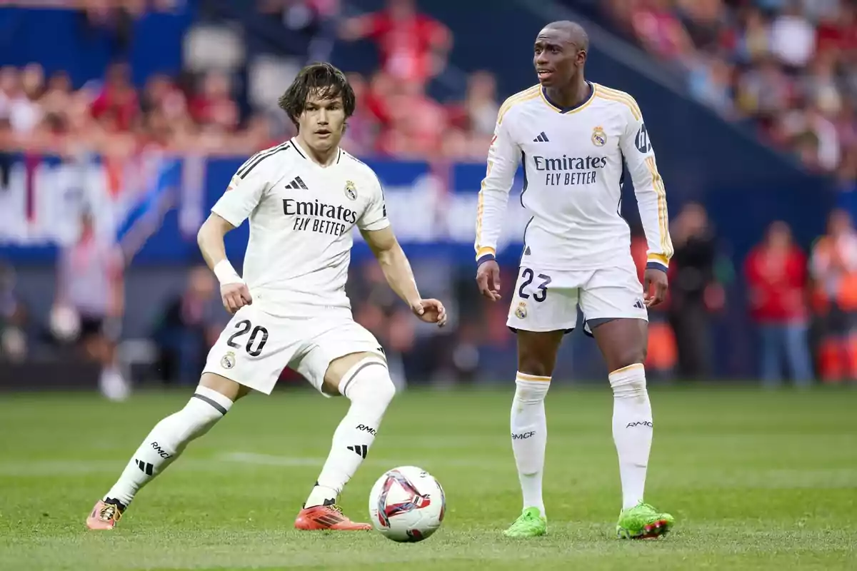 Dos jugadors de futbol del Reial Madrid al camp durant un partit, un d'ells està controlant la pilota mentre l'altre observa. Dos jugadors de futbol del Reial Madrid al camp durant un partit, un d'ells està controlant la pilota mentre l'altre observa.