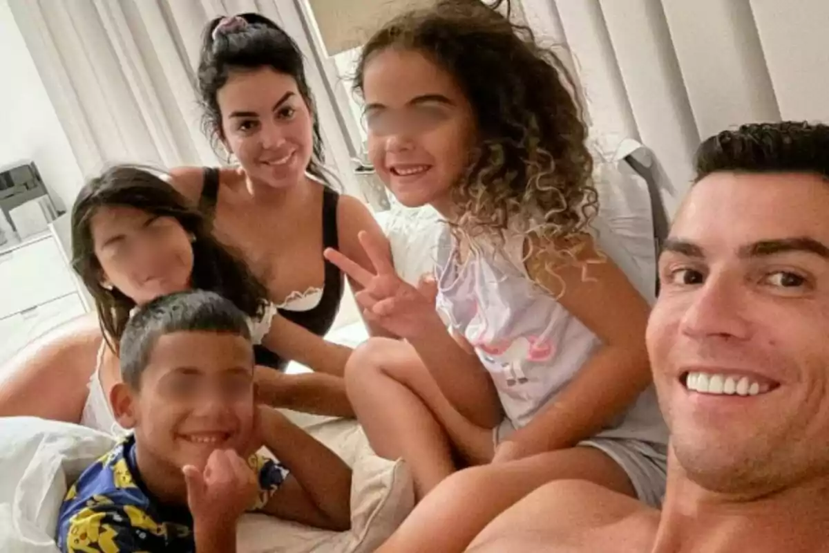 Georgina Rodríguez, Cristiano Ronaldo, and their three children pose smiling while taking a selfie in a room. Georgina Rodríguez, Cristiano Ronaldo, and their three children pose smiling while taking a selfie in a room.