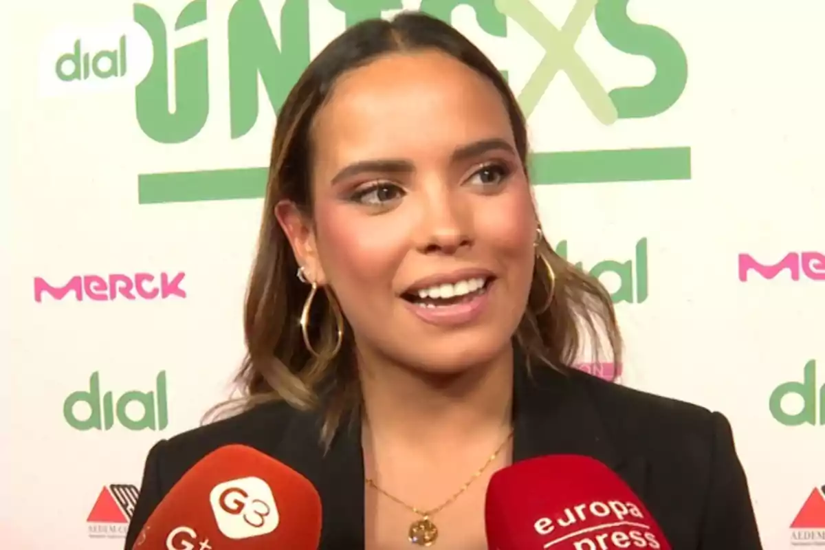 Gloria Camila somrient durant una entrevista en un esdeveniment amb micròfons de diferents mitjans davant seu i un fons amb logotips de patrocinadors.