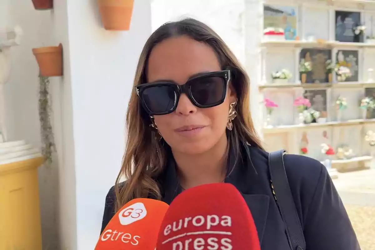 Gloria Camila amb ulleres de sol negres i jaqueta fosca respon preguntes davant de micròfons de premsa en un cementiri Gloria Camila amb ulleres de sol negres i jaqueta fosca respon preguntes davant de micròfons de premsa en un cementiri