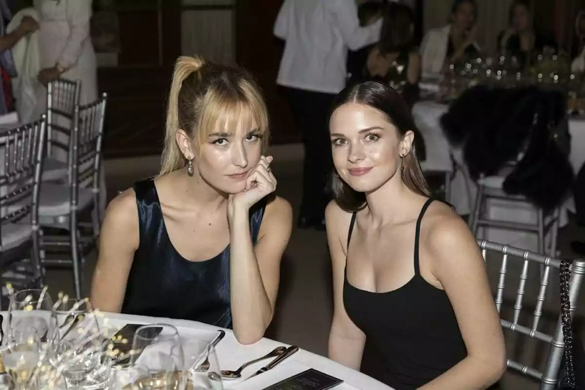 Grace Johnson i Stella del Carmen Banderas assegudes en una taula elegantment decorada en un esdeveniment formal, envoltades de copes i coberts, amb altres persones al fons. Grace Johnson i Stella del Carmen Banderas assegudes en una taula elegantment decorada en un esdeveniment formal, envoltades de copes i coberts, amb altres persones al fons.