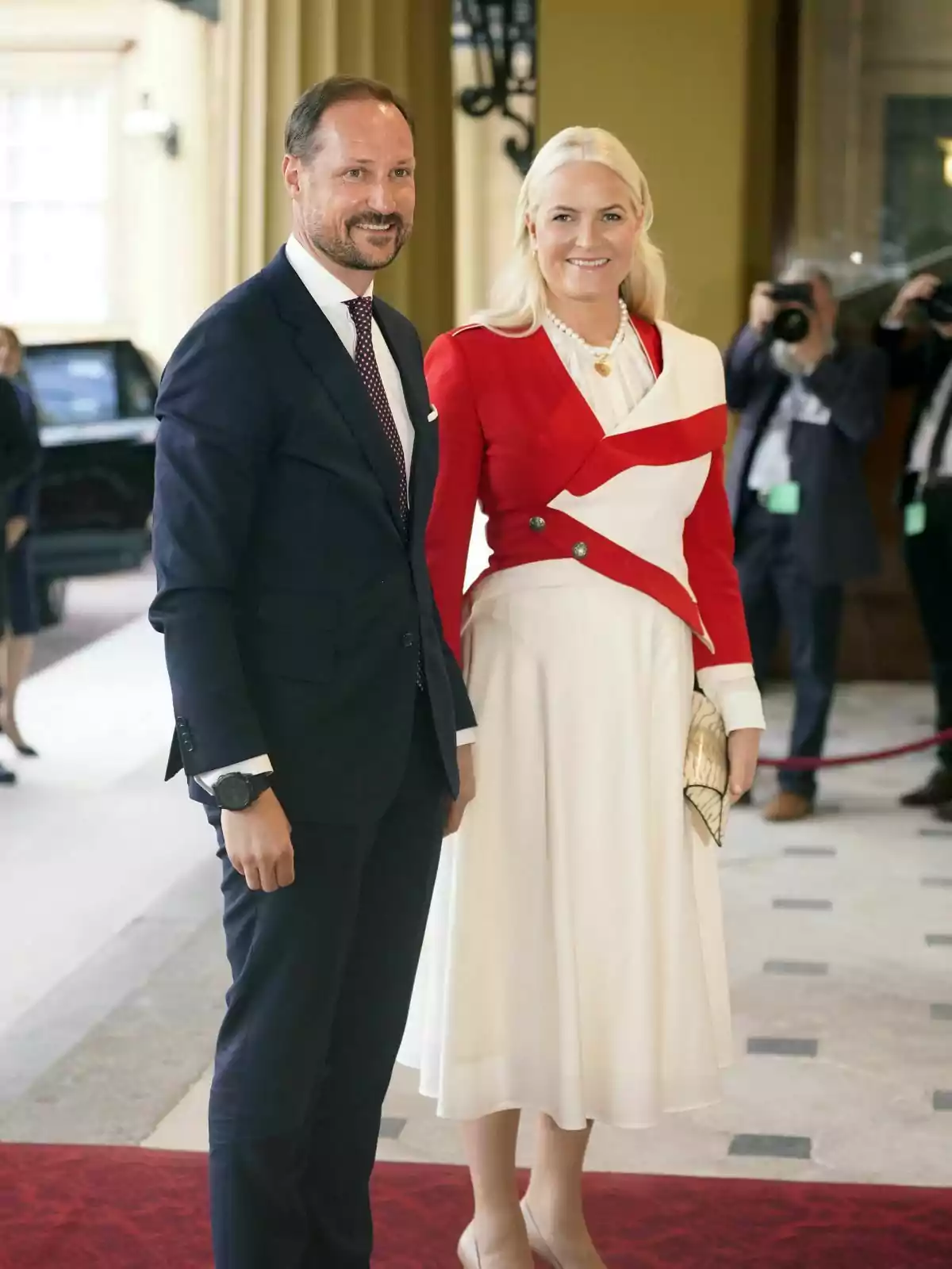 Haakon de Noruega i Mette-Marit elegantment vestits posen per a una foto en un esdeveniment formal, amb Haakon en vestit fosc i Mette-Marit amb un vestit blanc i jaqueta vermella, mentre fotògrafs capturen el moment al fons.