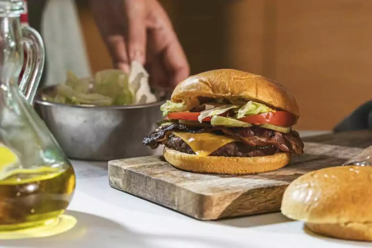 Hamburguesa amb formatge, bacó, enciam i tomàquet sobre una taula de fusta amb una mà al fons preparant ingredients Hamburguesa amb formatge, bacó, enciam i tomàquet sobre una taula de fusta amb una mà al fons preparant ingredients