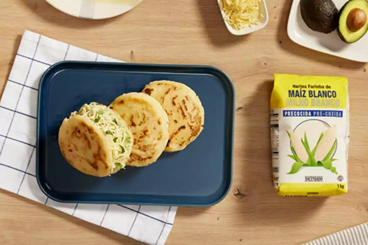 Tres arepas sobre un plat blau acompanyades d'ingredients com alvocat, formatge ratllat i farina de blat de moro blanc. Tres arepas sobre un plat blau acompanyades d'ingredients com alvocat, formatge ratllat i farina de blat de moro blanc.