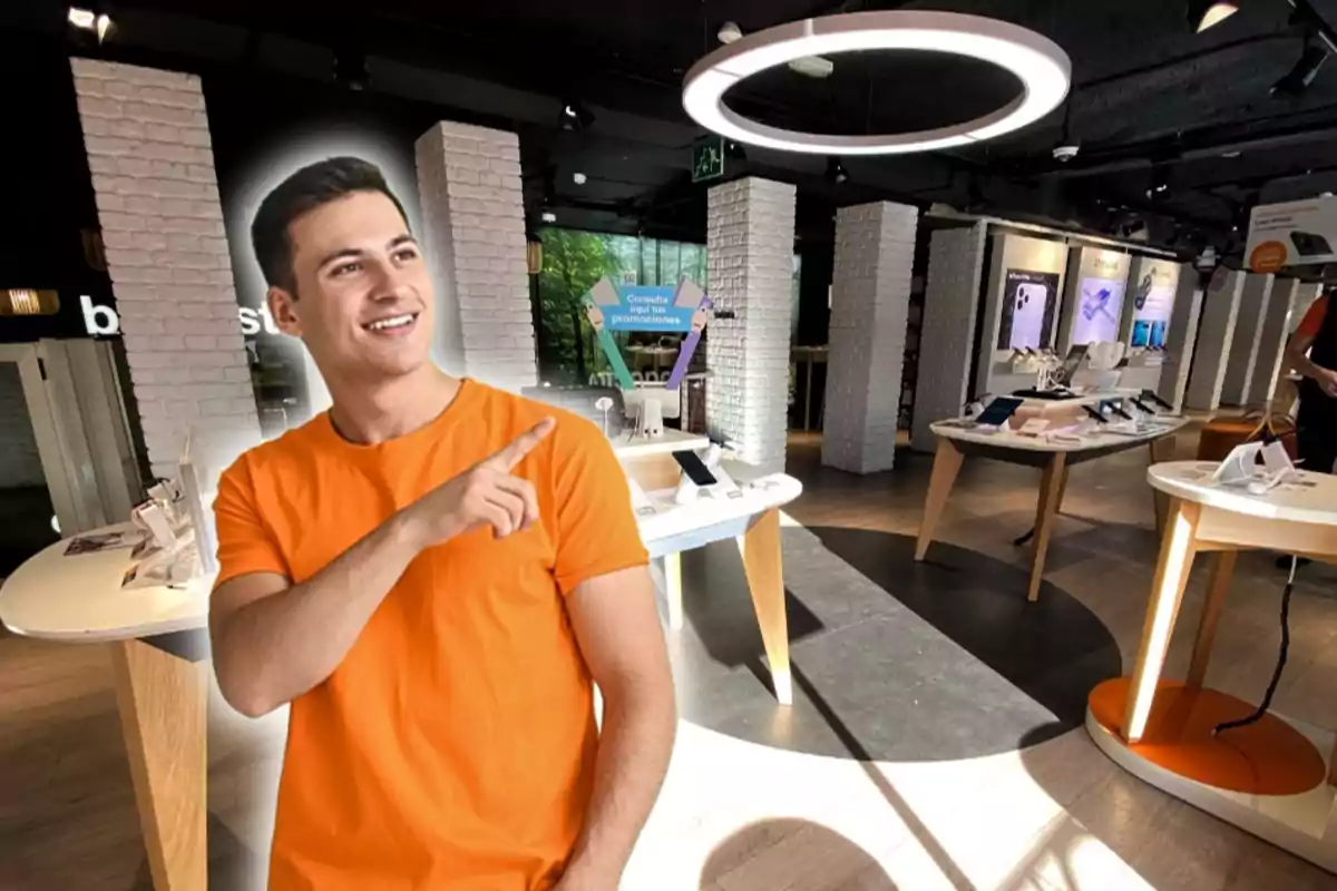 Un home amb samarreta taronja està somrient i assenyalant amb el dit en una botiga de tecnologia moderna amb taules que exhibeixen dispositius electrònics.