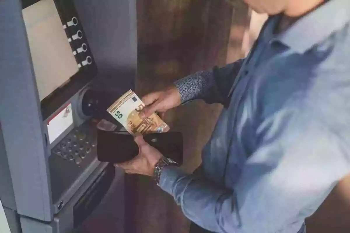 Home davant d'un caixer automàtic amb una cartera i bitllets a les mans