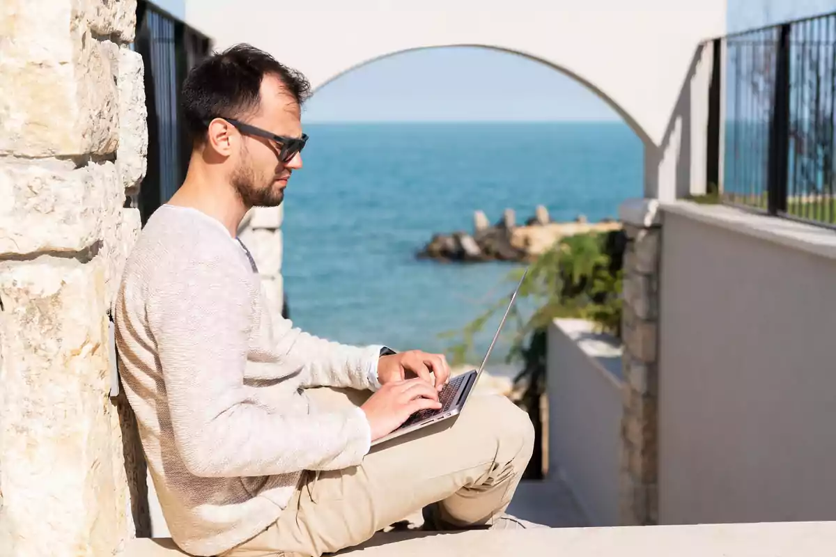 Hombre con gafas de sol usando una laptop mientras está sentado al aire libre junto a una pared de piedra con vista al mar.