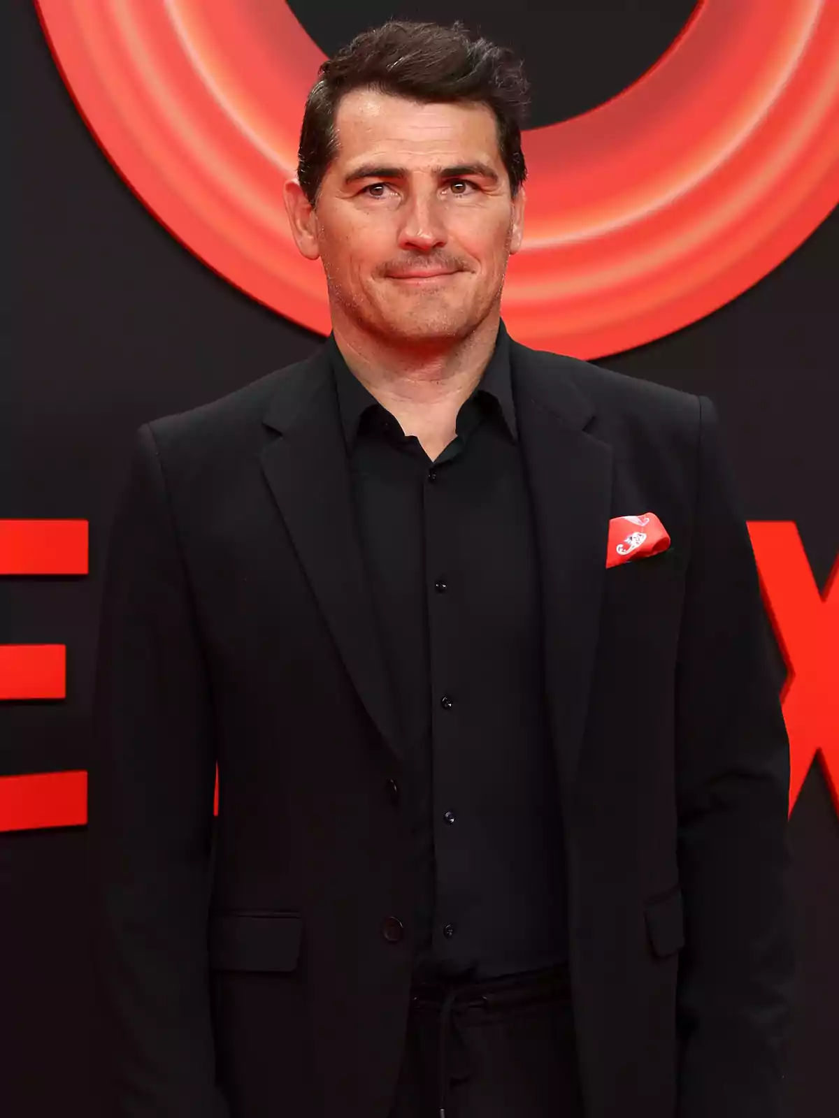 Íker Casillas vestint un vestit negre amb camisa fosca i mocador vermell a la solapa, posant davant d’un fons negre amb detalls vermells. Íker Casillas vestint un vestit negre amb camisa fosca i mocador vermell a la solapa, posant davant d’un fons negre amb detalls vermells.