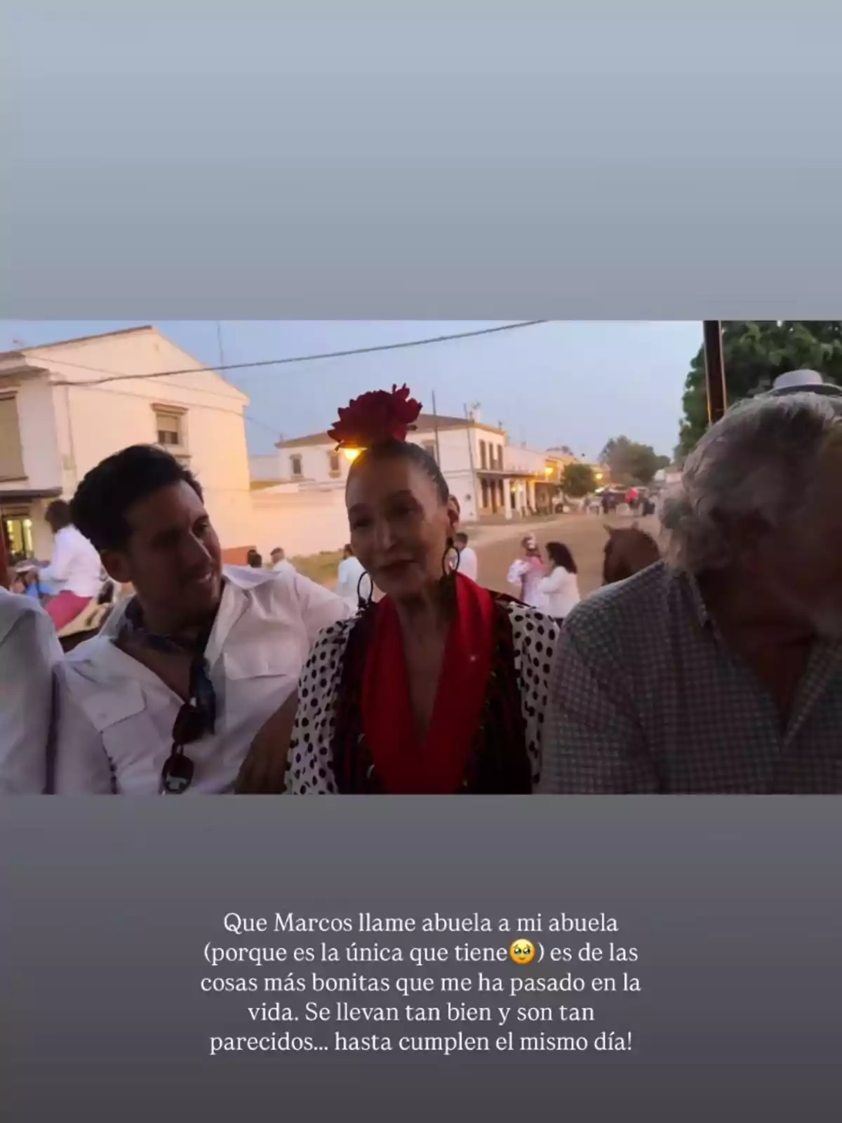 Tres persones assegudes juntes a l’aire lliure, una dona al centre porta una flor vermella al cap i vesteix roba tradicional, mentre els homes al seu costat l’acompanyen somrients; al fons es veuen cases i més persones al carrer. Tres persones assegudes juntes a l’aire lliure, una dona al centre porta una flor vermella al cap i vesteix roba tradicional, mentre els homes al seu costat l’acompanyen somrients; al fons es veuen cases i més persones al carrer.