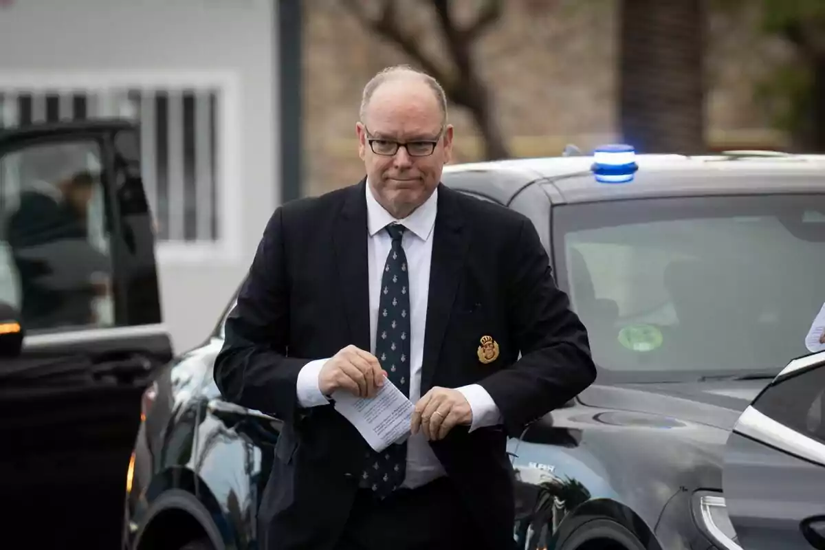 A man in a dark suit and tie holds a paper while standing in front of a car with a blue light on the roof. A man in a dark suit and tie holds a paper while standing in front of a car with a blue light on the roof.