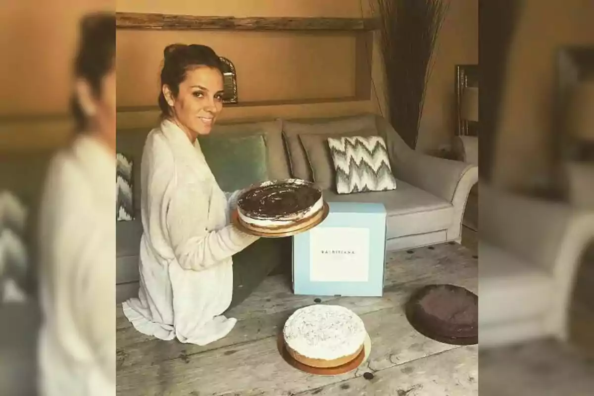 A person sitting on the floor of a room holds a cake while smiling at the camera, with a blue box and other cakes on a wooden table in front of a sofa. A person sitting on the floor of a room holds a cake while smiling at the camera, with a blue box and other cakes on a wooden table in front of a sofa.