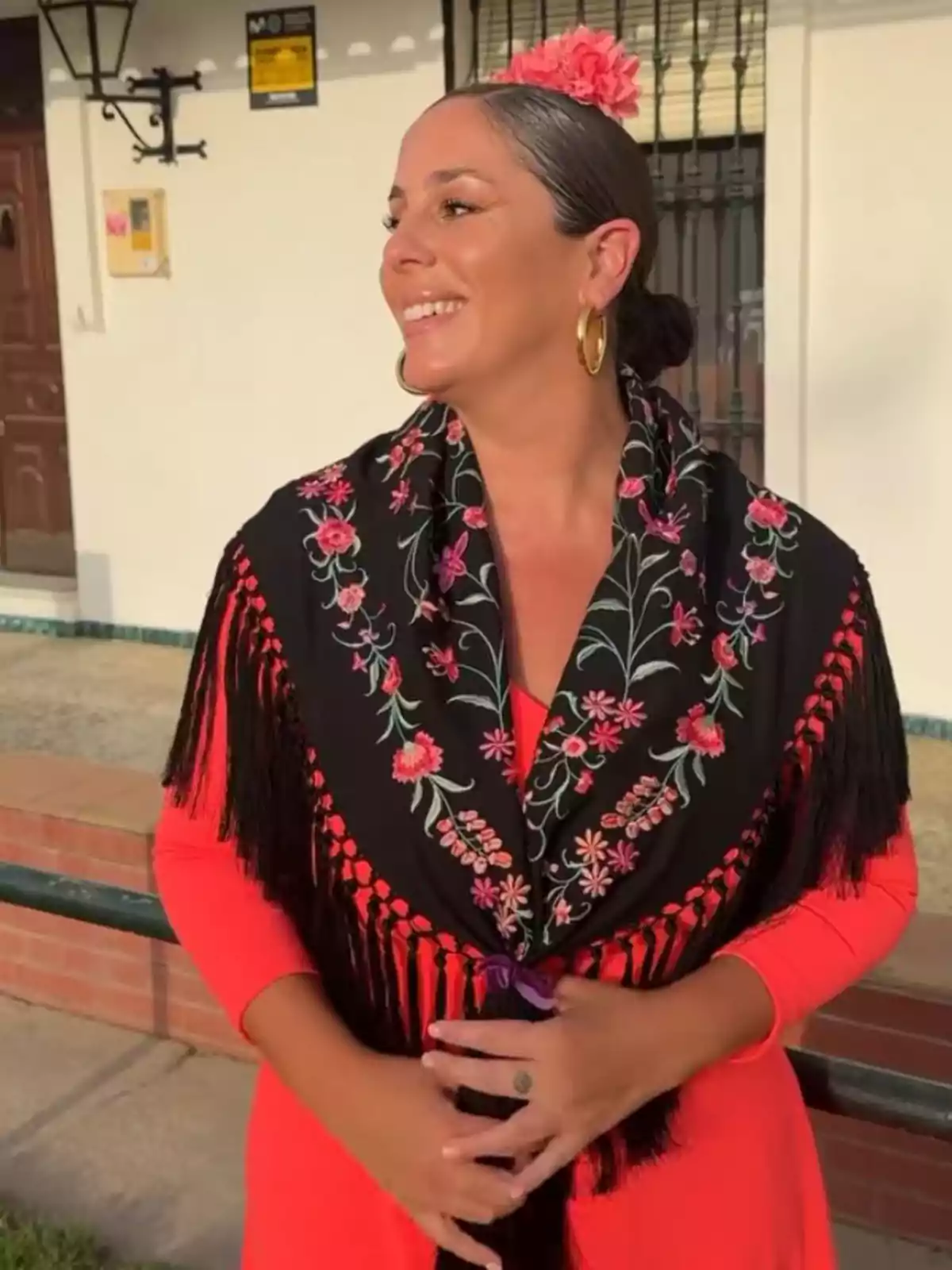 Dona somrient vestida amb vestit tradicional flamenc de color vermell i mantó negre brodat amb flors, porta una flor rosa als cabells i arracades daurades, posant a l’aire lliure davant d’una casa. Dona somrient vestida amb vestit tradicional flamenc de color vermell i mantó negre brodat amb flors, porta una flor rosa als cabells i arracades daurades, posant a l’aire lliure davant d’una casa.