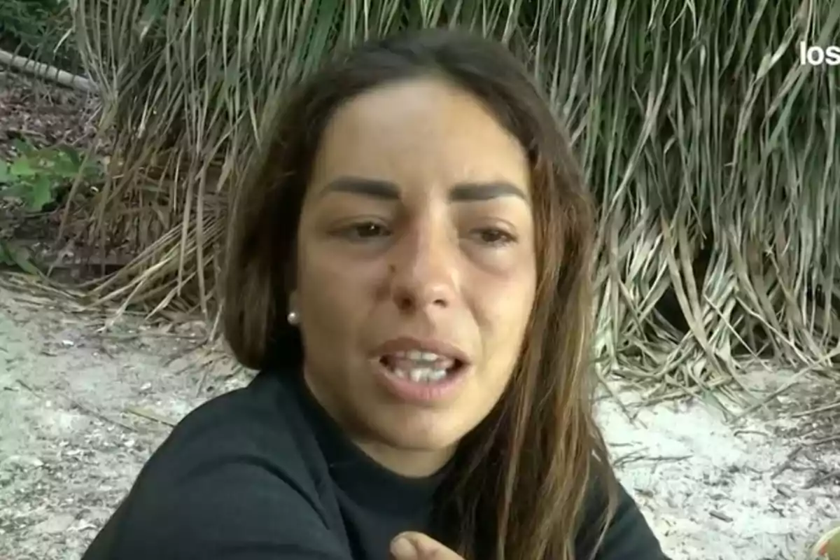 Una dona amb expressió seriosa està asseguda en una platja amb vegetació al fons. Una dona amb expressió seriosa està asseguda en una platja amb vegetació al fons.