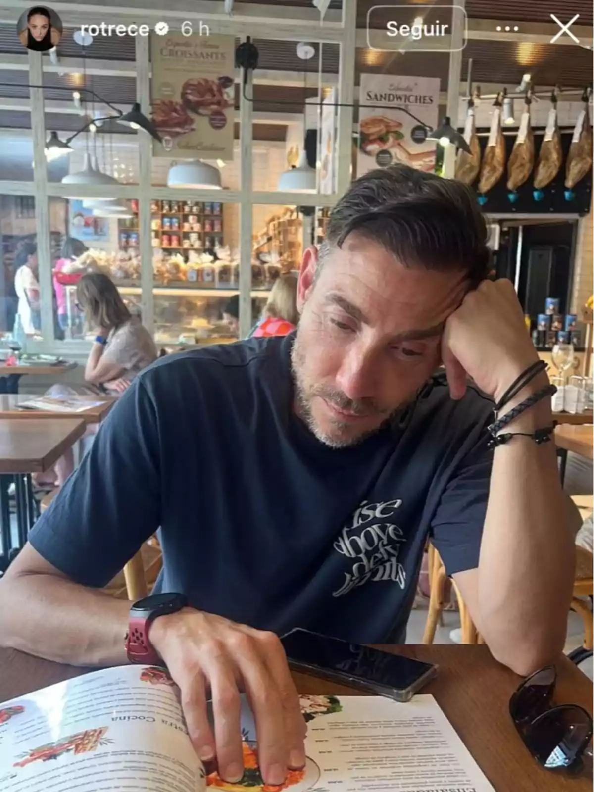 Home assegut en una cafeteria mirant el menú amb expressió pensativa, porta una samarreta fosca i té un rellotge vermell al canell, sobre la taula hi ha un parell d’ulleres de sol i un telèfon mòbil. Home assegut en una cafeteria mirant el menú amb expressió pensativa, porta una samarreta fosca i té un rellotge vermell al canell, sobre la taula hi ha un parell d’ulleres de sol i un telèfon mòbil.