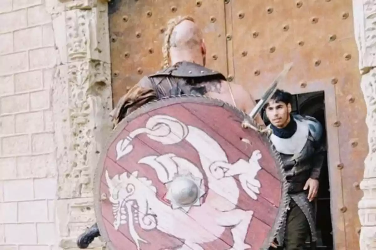 Un guerrer amb escut decorat s'enfronta a un home que surt d'una porta de fusta. Un guerrer amb escut decorat s'enfronta a un home que surt d'una porta de fusta.
