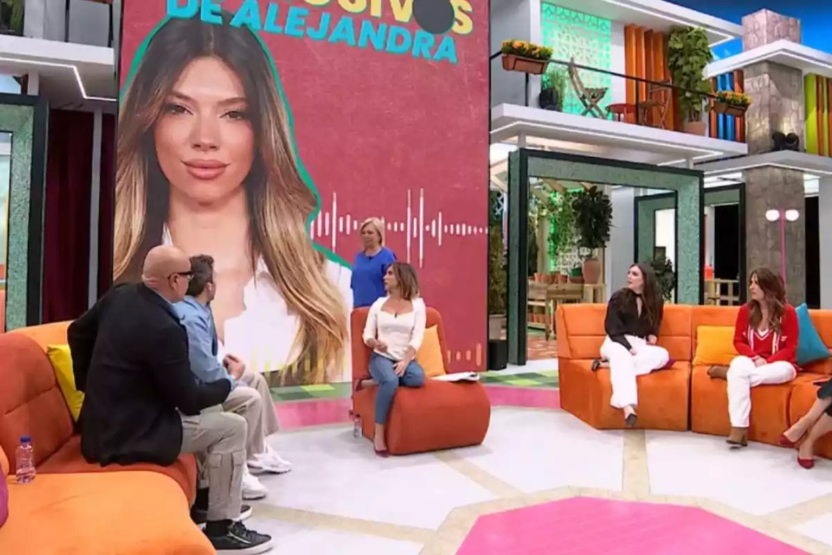 Un grup de persones està assegut en un plató de televisió acolorit, amb un gran cartell de fons que mostra la imatge d'una dona i el text Un grup de persones està assegut en un plató de televisió acolorit, amb un gran cartell de fons que mostra la imatge d'una dona i el text