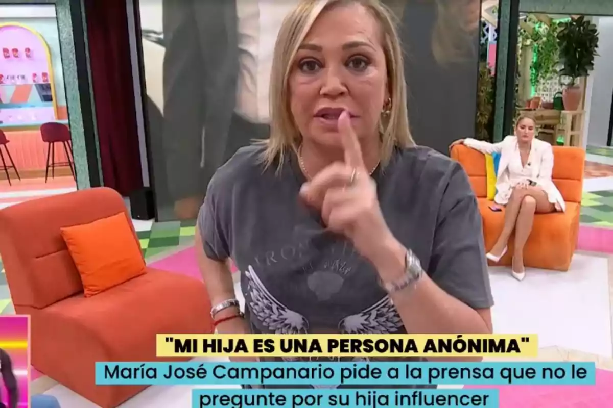 Una dona en un plató de televisió fa un gest amb el dit mentre una altra persona està asseguda en un sofà taronja al fons. Una dona en un plató de televisió fa un gest amb el dit mentre una altra persona està asseguda en un sofà taronja al fons.