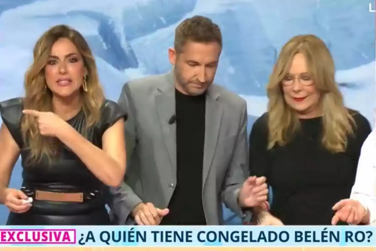 Tres persones són en un plató de televisió, una dona a l'esquerra assenyala cap a un costat mentre un home al centre i una altra dona a la dreta miren cap avall, a la part inferior de la imatge hi ha un text que diu Tres persones són en un plató de televisió, una dona a l'esquerra assenyala cap a un costat mentre un home al centre i una altra dona a la dreta miren cap avall, a la part inferior de la imatge hi ha un text que diu