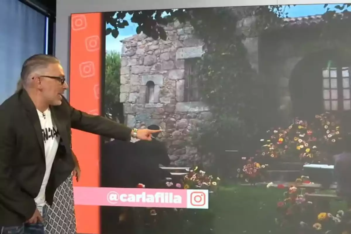 Home amb ulleres i jaqueta fosca assenyalant una pantalla gran que mostra una casa de pedra envoltada de flors i vegetació