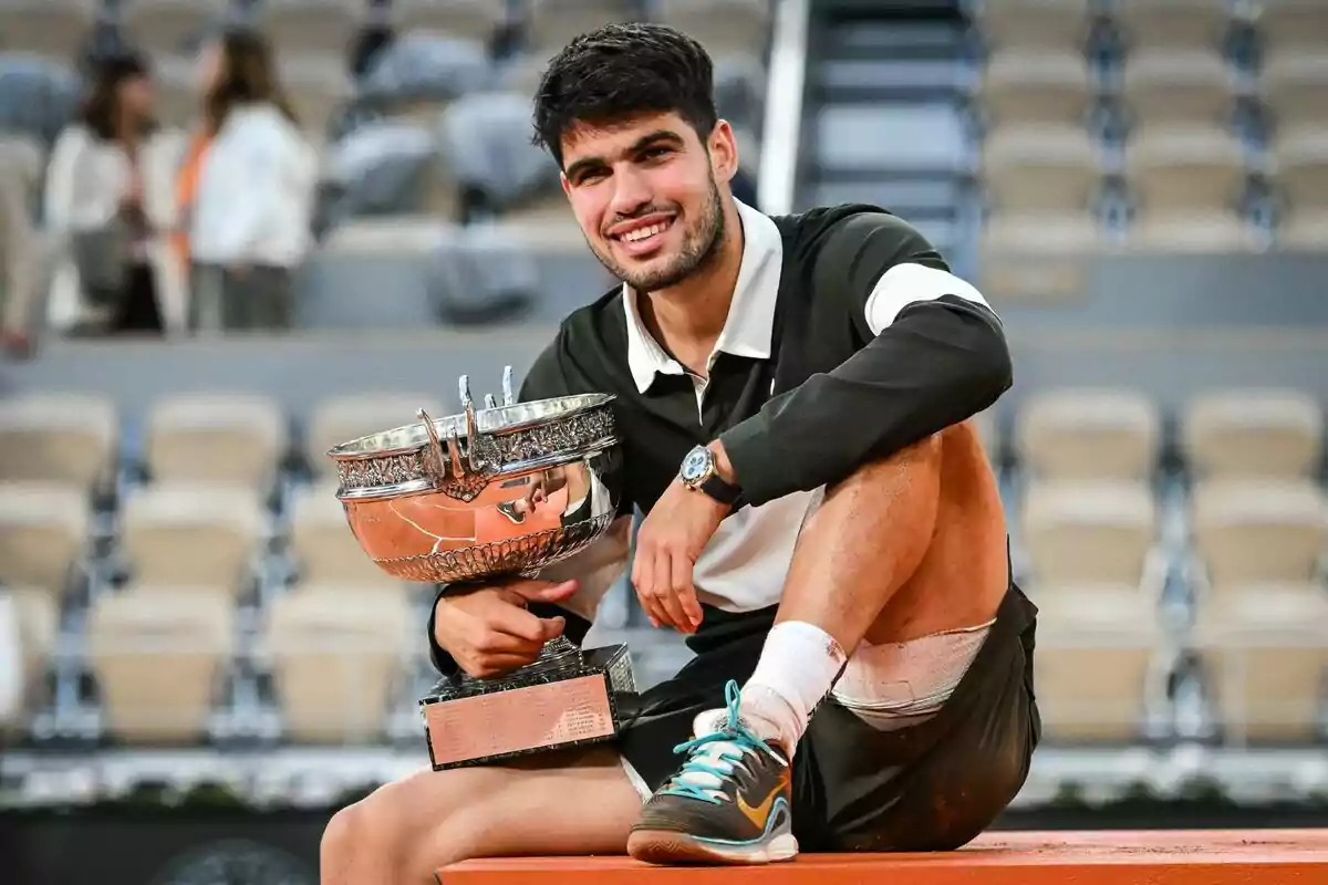 Smiling young tennis player sitting on a court holding a large silver trophy with empty stands in the background Smiling young tennis player sitting on a court holding a large silver trophy with empty stands in the background
