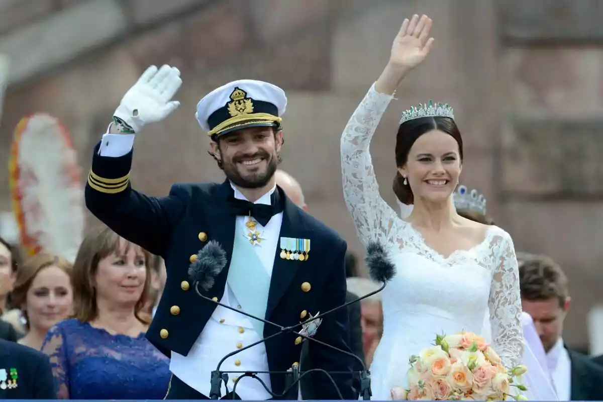 A couple dressed in formal attire smiles and waves while the woman holds a bouquet of flowers. A couple dressed in formal attire smiles and waves while the woman holds a bouquet of flowers.