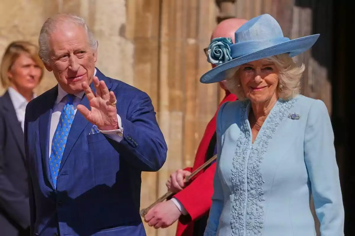 Two elegantly dressed elderly people, one wearing a blue suit with a matching tie, wave as they walk outdoors, the other person is wearing a light blue dress with a hat adorned with a flower. Two elegantly dressed elderly people, one wearing a blue suit with a matching tie, wave as they walk outdoors, the other person is wearing a light blue dress with a hat adorned with a flower.