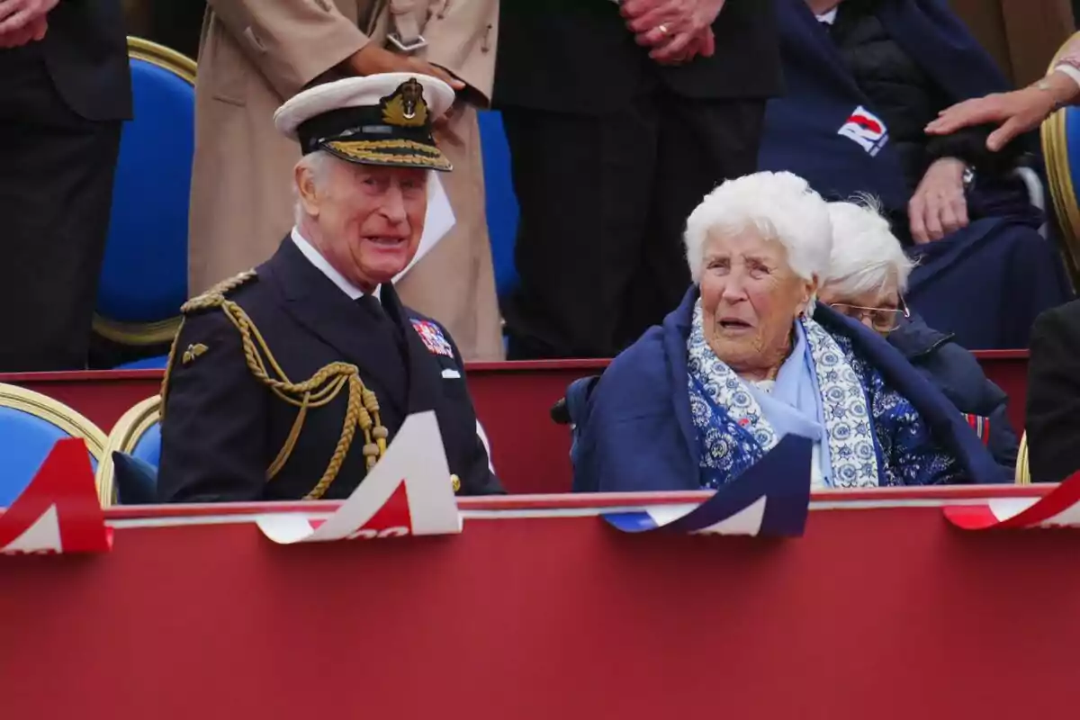 Un hombre con uniforme naval y una mujer mayor están sentados en una tribuna decorada con banderas. Un hombre con uniforme naval y una mujer mayor están sentados en una tribuna decorada con banderas.