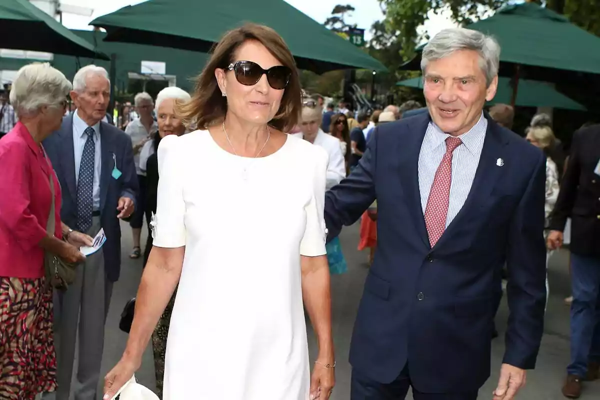 A well-dressed couple walks outdoors surrounded by other people at a social event.