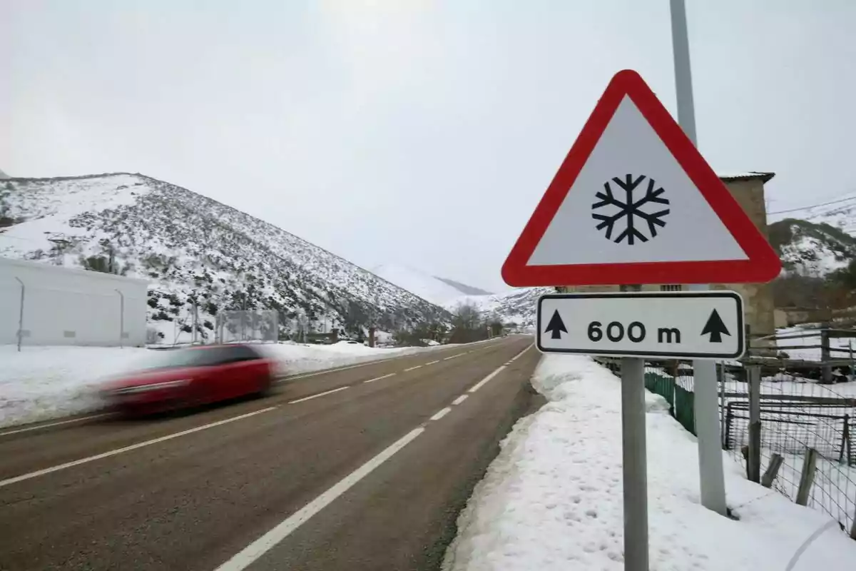 Un coche rojo pasa rápidamente por una carretera nevada con una señal de advertencia de nieve a 600 metros.