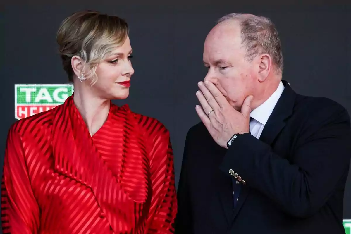 A woman in a red dress looks at a man in a dark suit who covers his mouth with his hand while speaking to her at a formal event. A woman in a red dress looks at a man in a dark suit who covers his mouth with his hand while speaking to her at a formal event.