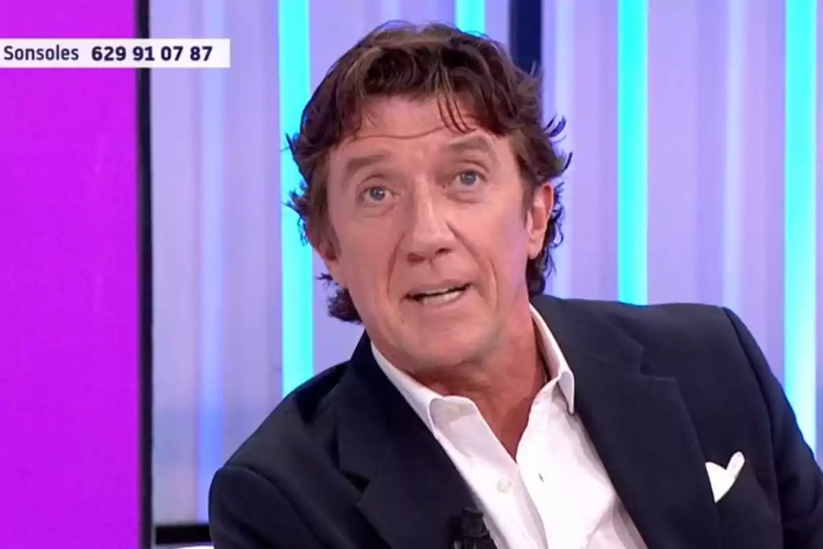 Home de cabells castanys i ulls clars amb jaqueta fosca i camisa blanca en un plató de televisió amb fons de llums blaves i liles Home de cabells castanys i ulls clars amb jaqueta fosca i camisa blanca en un plató de televisió amb fons de llums blaves i liles