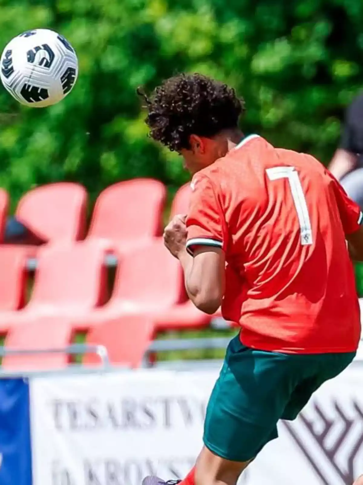 Jugador de futbol amb samarreta vermella i número set remata de cap una pilota en un camp amb seients vermells i fons verd. Jugador de futbol amb samarreta vermella i número set remata de cap una pilota en un camp amb seients vermells i fons verd.