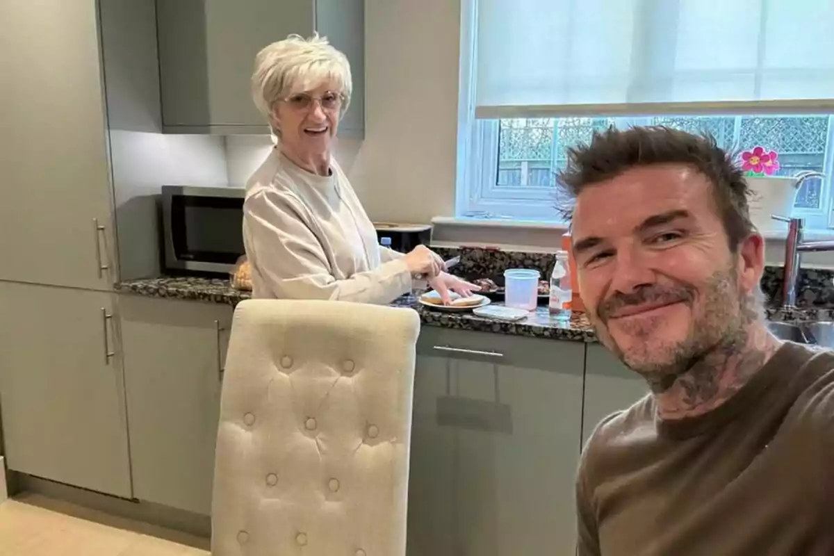 An elderly person is in the kitchen preparing something while another person smiles at the camera. An elderly person is in the kitchen preparing something while another person smiles at the camera.