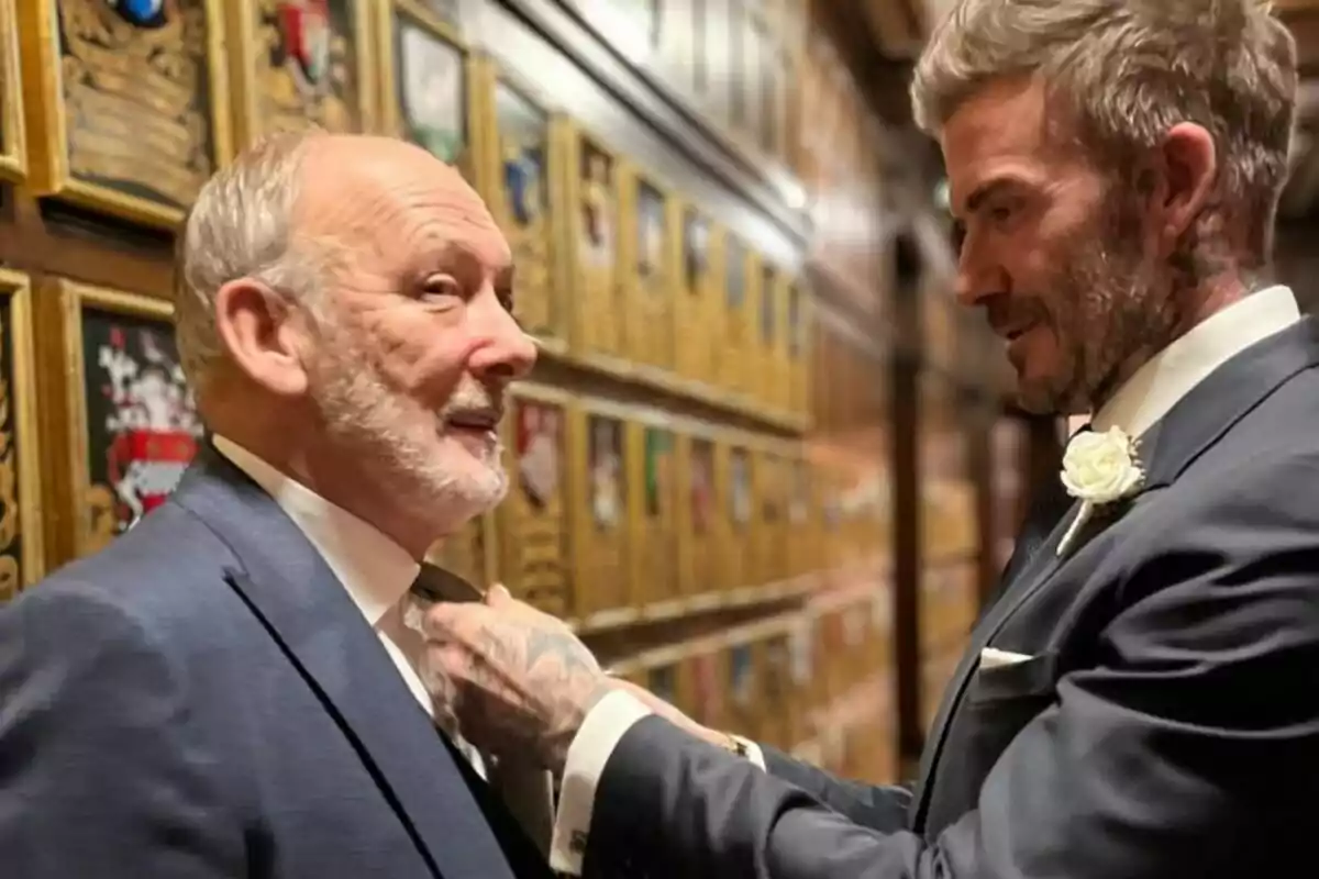 A man with a beard and gray hair is being helped with his tie by another man in a dark suit with a flower on the lapel, in a hallway decorated with golden plaques. A man with a beard and gray hair is being helped with his tie by another man in a dark suit with a flower on the lapel, in a hallway decorated with golden plaques.