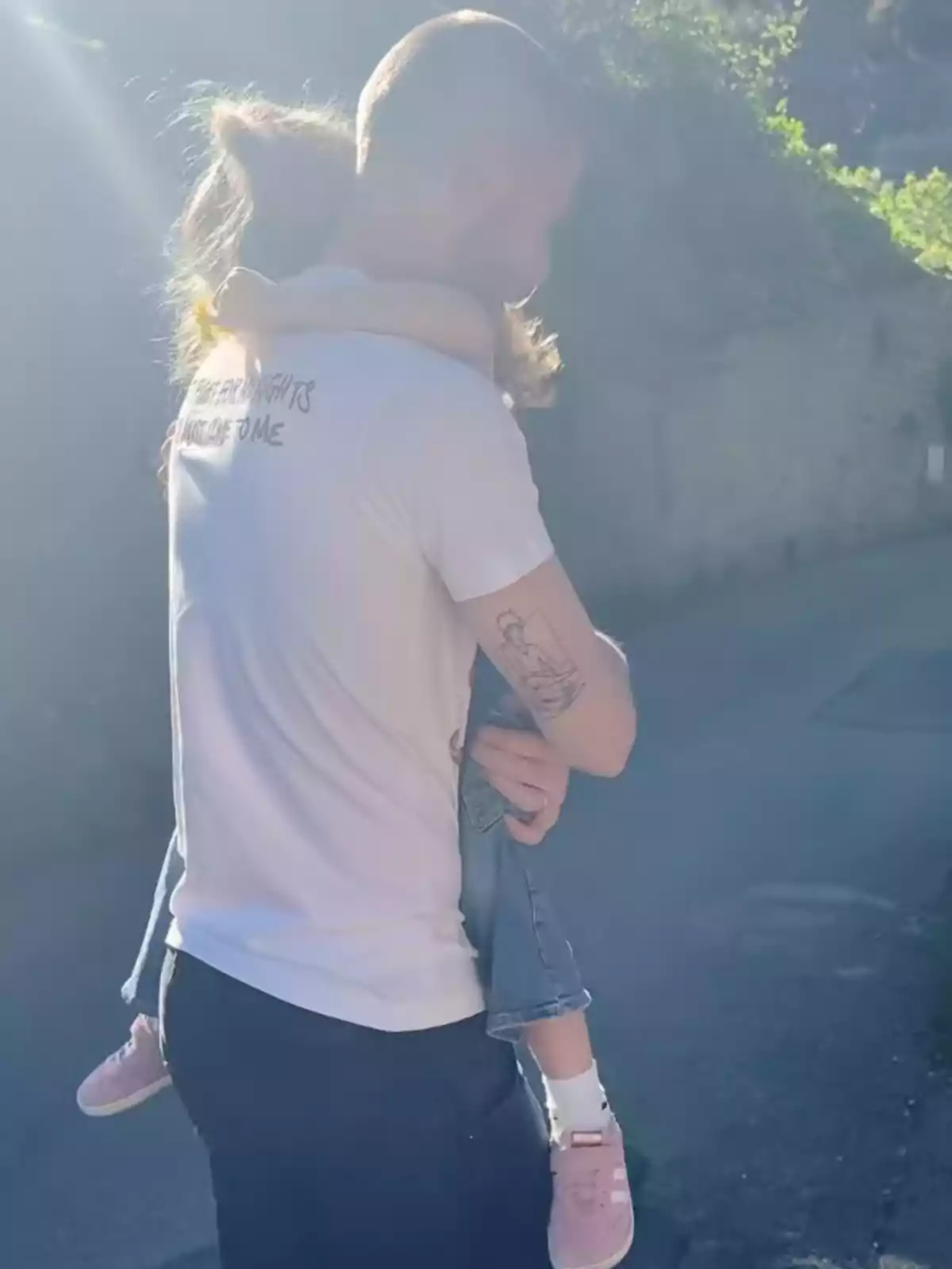 Un home amb samarreta blanca i tatuatge al braç sosté una nena amb sabatilles roses en un camí il·luminat pel sol. Un home amb samarreta blanca i tatuatge al braç sosté una nena amb sabatilles roses en un camí il·luminat pel sol.