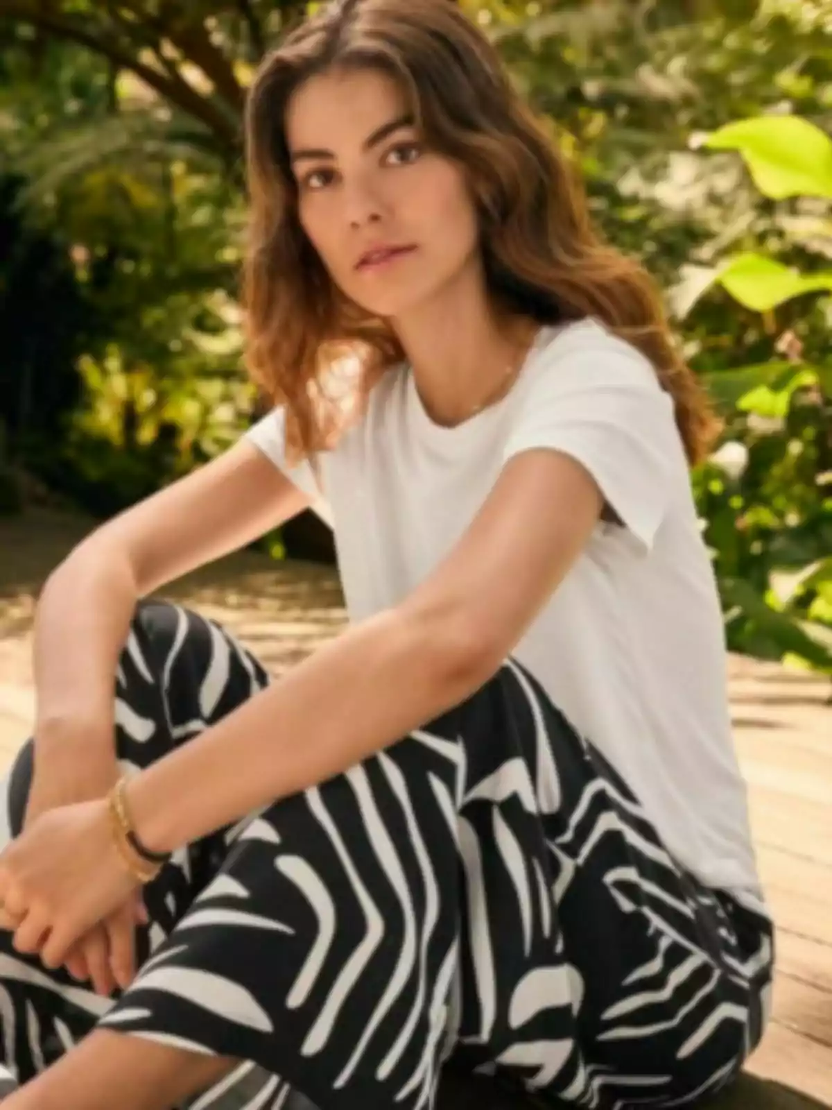 Una persona amb cabell castany i ondulat, vestida amb una samarreta blanca i pantalons amb estampat de ratlles de Lidl, està asseguda a l'aire lliure en un entorn natural. Una persona amb cabell castany i ondulat, vestida amb una samarreta blanca i pantalons amb estampat de ratlles de Lidl, està asseguda a l'aire lliure en un entorn natural.