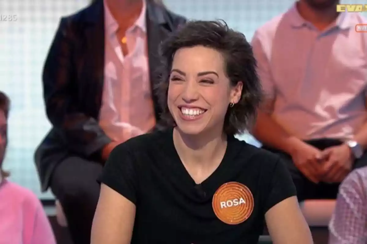 Dona somrient amb samarreta negra i un gafet taronja que diu Rosa participant en un programa de televisió amb públic al fons Dona somrient amb samarreta negra i un gafet taronja que diu Rosa participant en un programa de televisió amb públic al fons