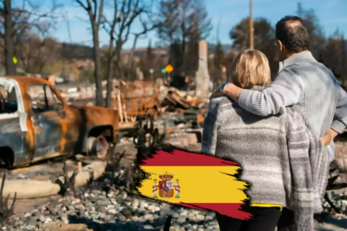 Una parella observa un paisatge devastat amb un cotxe cremat i arbres sense fulles mentre una bandera d?Espanya apareix a la part inferior de la imatge.