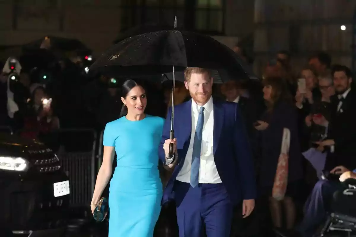 An elegant couple walks in the rain, sharing a black umbrella while being photographed by a crowd of people. An elegant couple walks in the rain, sharing a black umbrella while being photographed by a crowd of people.