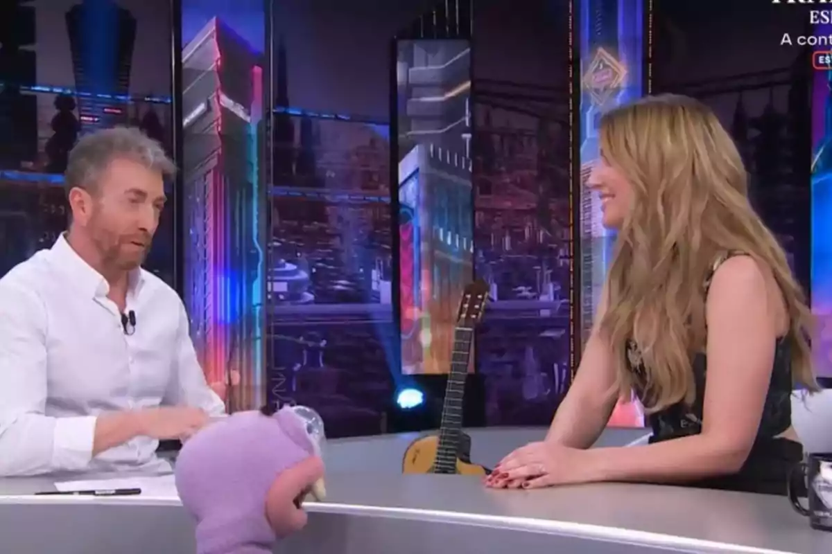 Un home i una dona conversen en un plató de televisió amb un fons acolorit i una guitarra recolzada darrere d'ells, mentre un personatge de peluix està en primer pla. Un home i una dona conversen en un plató de televisió amb un fons acolorit i una guitarra recolzada darrere d'ells, mentre un personatge de peluix està en primer pla.
