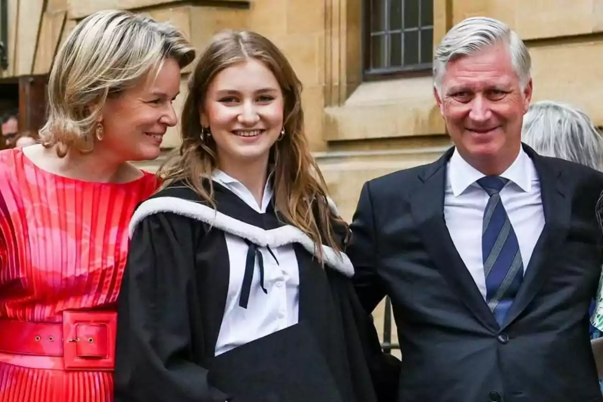 A young woman in a graduation gown smiles next to two older people, a woman in a red dress and a man in a dark suit, in a classical architectural setting. A young woman in a graduation gown smiles next to two older people, a woman in a red dress and a man in a dark suit, in a classical architectural setting.