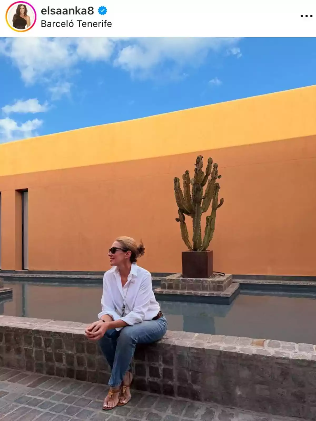 Elsa Anka amb ulleres de sol i camisa blanca està asseguda en una vora de pedra al costat d'un estany, amb un cactus i una paret de color taronja de fons sota un cel blau.