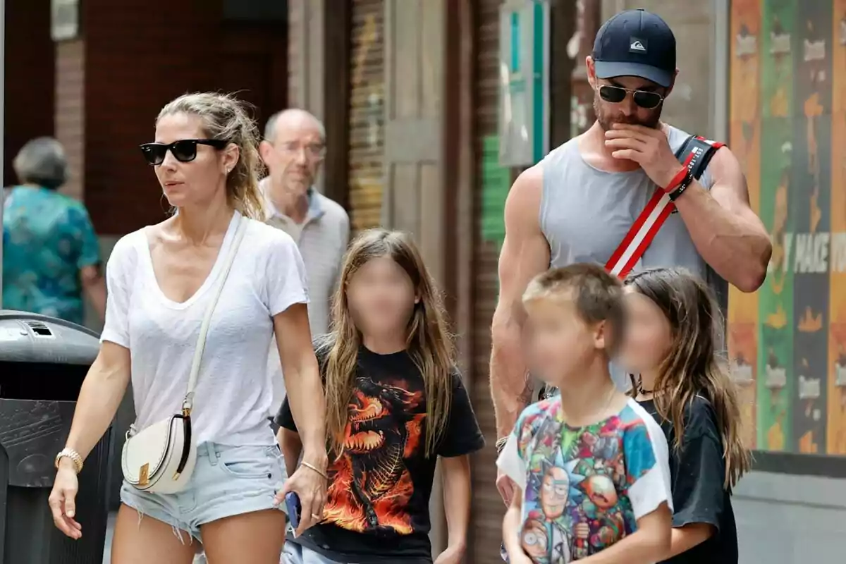 Una pareja camina por la calle acompañada de tres niños, todos llevan gafas de sol y ropa casual, mientras un hombre mayor camina detrás de ellos. Una pareja camina por la calle acompañada de tres niños, todos llevan gafas de sol y ropa casual, mientras un hombre mayor camina detrás de ellos.