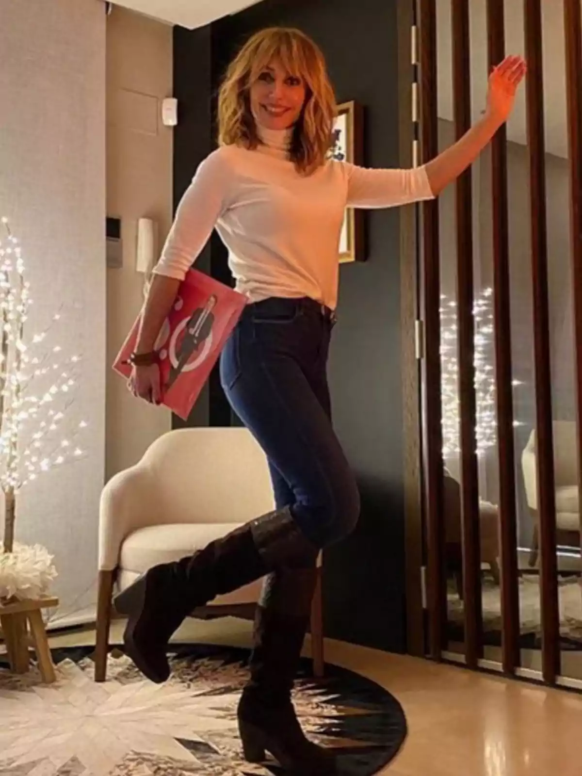 Una persona amb cabell ros i solt, vestida amb un jersei blanc i texans foscos, sosté un llibre vermell mentre posa dreta en una habitació decorada amb llums i mobles moderns. Una persona amb cabell ros i solt, vestida amb un jersei blanc i texans foscos, sosté un llibre vermell mentre posa dreta en una habitació decorada amb llums i mobles moderns.