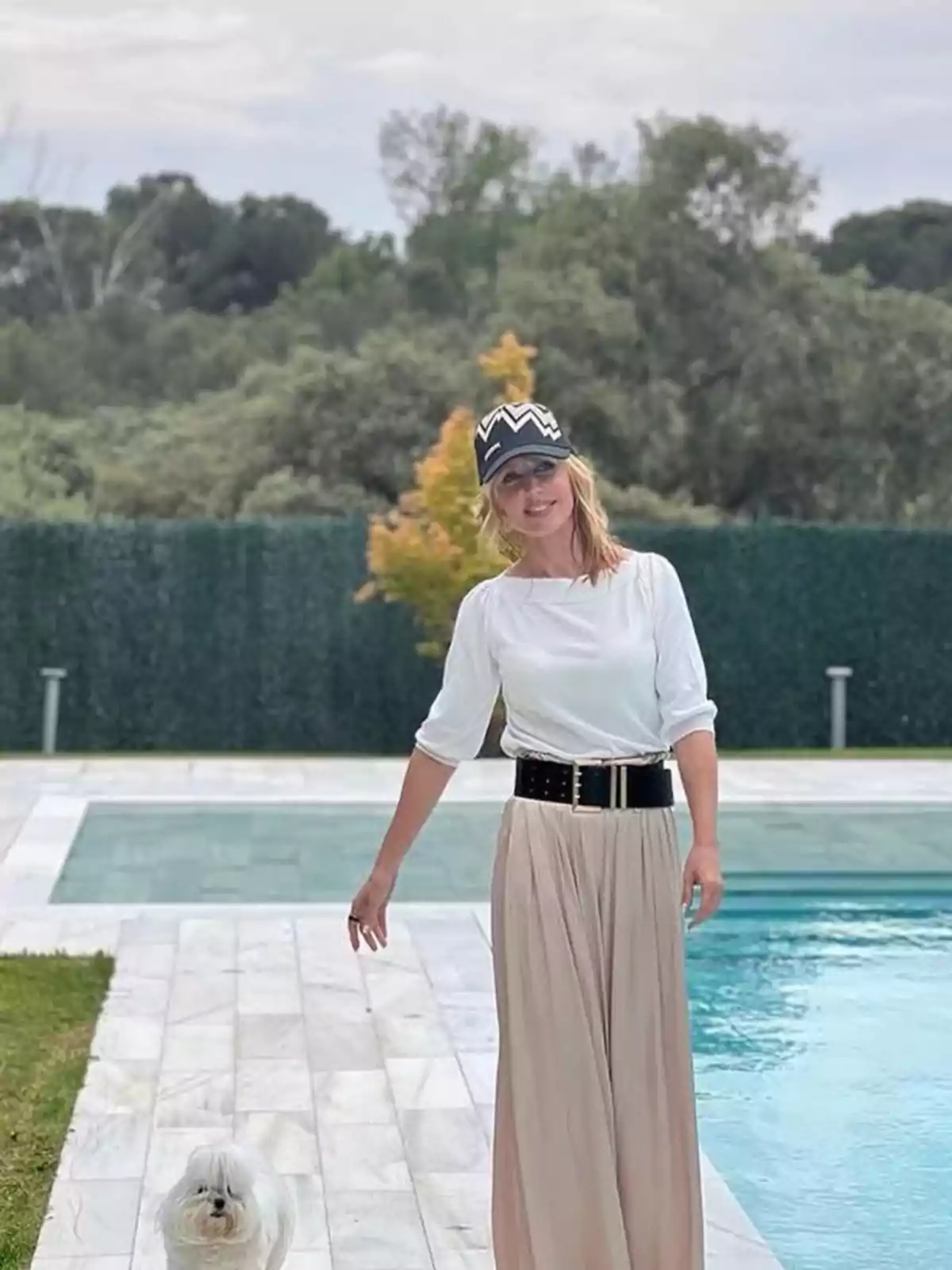 Una persona amb barret i faldilla llarga està dreta al costat d'una piscina, acompanyada d'un gos petit, amb un fons d'arbres i bardisses. Una persona amb barret i faldilla llarga està dreta al costat d'una piscina, acompanyada d'un gos petit, amb un fons d'arbres i bardisses.