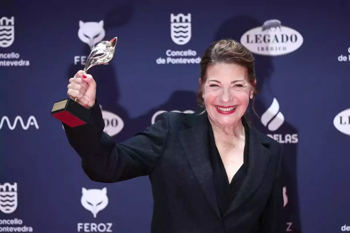 Una persona sonriente sostiene un trofeo en un evento con un fondo de logotipos. Una persona sonriente sostiene un trofeo en un evento con un fondo de logotipos.
